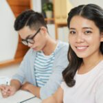 Pretty modern Asian woman at workplace with young male coworker near smiling at camera