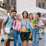 Laughing asian boy in glasses and shorts embracing charming blonde girls in front of outdoor cafe. Joyful students came to open-air restaurant to celebrate end of exams